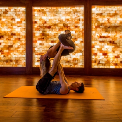 A Man And Woman In A Yoga Pose On An Orange Yoga Mat On A Wooden Floor In Front Of A Backlit Orange Himalayan Salt Stone Wall.