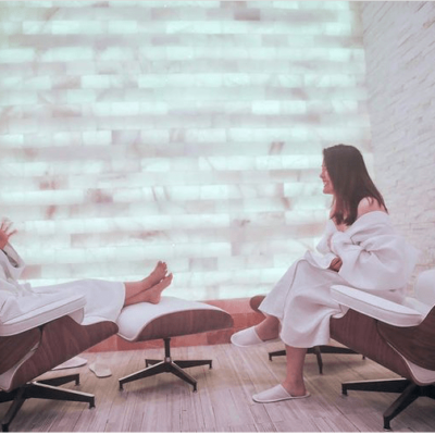 Two Women In White Robes Sitting In Two Chairs With Foot Stands In A Room With A White Backlit Salt Stone Wall.