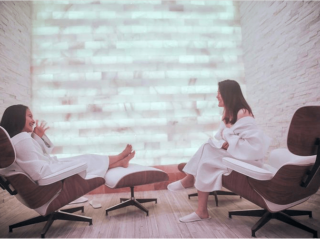 Two Women Sitting In White Robes In A Salt Therapy Room In Front Of A White Himalayan Salt Stone Wall Backlit With White Lighting.