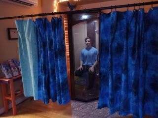 Man In Blue Shirt Sitting In A Wooden And Glass Salt Booth With Two Dark Blue Tied Dyed Curtains And One Light Blue Curtain On A Rail At The Infinity More Healing In Arlington Virginia