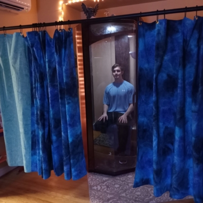 Man in blue shirt sitting in a wooden and glass salt booth with two dark blue tied dyed curtains and one light blue curtain on a rail at the Infinity More Healing in Arlington Virginia