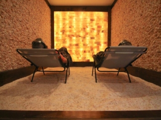Two People With Headsets Reclined In Chairs On A Salt-Covered Floor With Salt Stone Walls And An Orange Backlit Himalayan Paneled Wall.