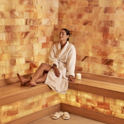 Woman in a white robe relaxing on a light brown wooden bench with Himalayan salt panel walls at the JW Marriott Turnberry - Aventura, Florida.