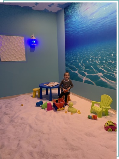 Young girl playing with toys on a salt-covered floor at a blue table in front of an ocean wallpaper at the Just Breathe Salt Spa in Hyannis, Massachusetts.