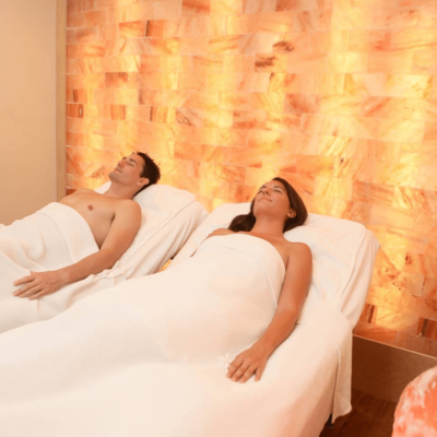 Man And Woman Relaxing On A Massage Table In Front Of A Backlit Salt Stone Wall With Orange Lighting.