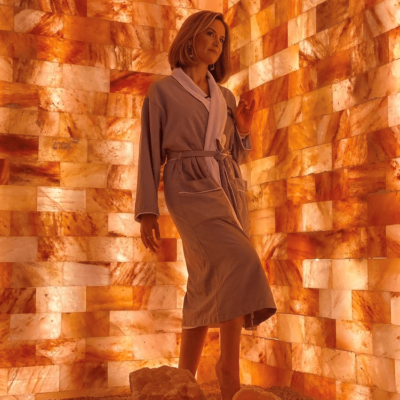 Woman In A Grey And White Robe Standing On A Salt-Covered Floor Leaning Against An Orange Backlit Salt Stone Wall.
