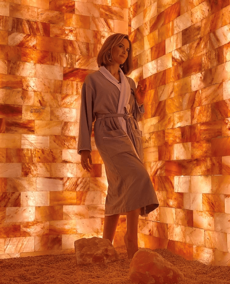 Woman In A Grey And White Robe Standing On A Salt-Covered Floor Leaning Against A Led Backlit Salt Panel Wall At The Kalahari - Round Rock, Texas