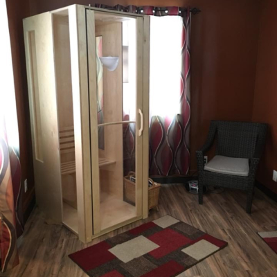 A light wooden and glass halotherapy booth in a red room with brown vinyl flooring and a brown chair in the corner at the Lifetime Wellness Chiropractic Center