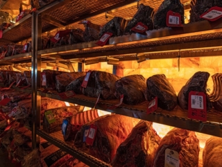 Five Shelves With Different Types Of Meat With Price Tags In Front Of A Himalayan Salt Paneled Wall Backlit By Orange Lighting At Meat On Ocean