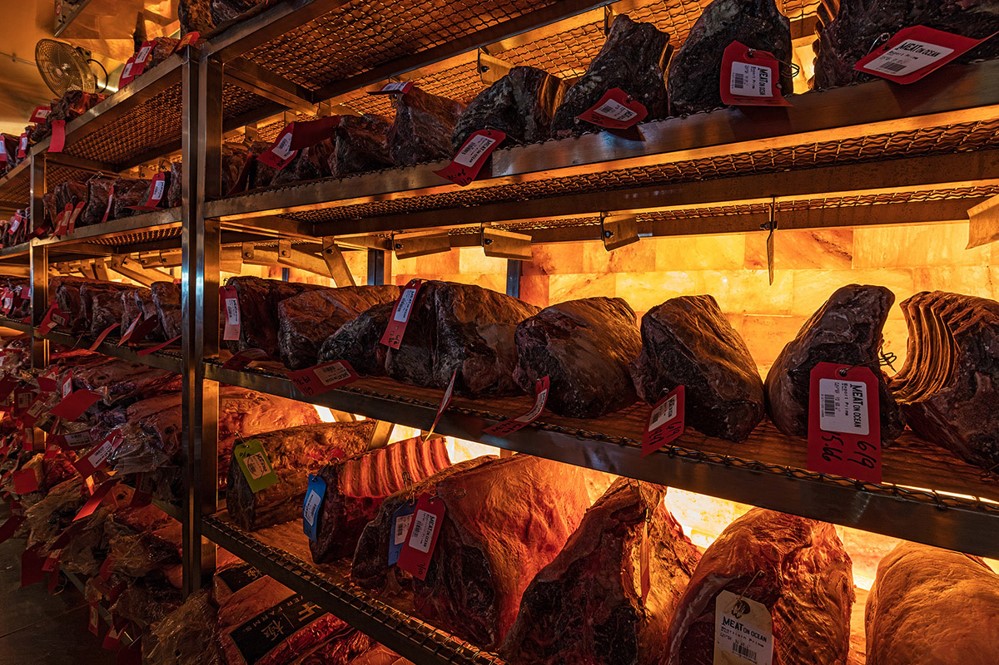 Five shelves with different types of meat with price tags in front of a Himalayan salt paneled wall backlit by orange lighting at Meat On Ocean