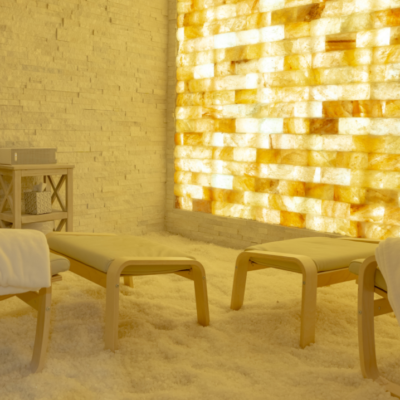 Two White Chairs With Foot Stools On A White Salt-Covered Floor With White Tiled Salt Walls And A Orange Backlit Salt Stone Wall.