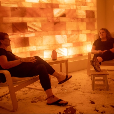 Two women sitting talking in a salt therapy room with a salt-covered floor and an orange backlit Himalayan salt stone wall.