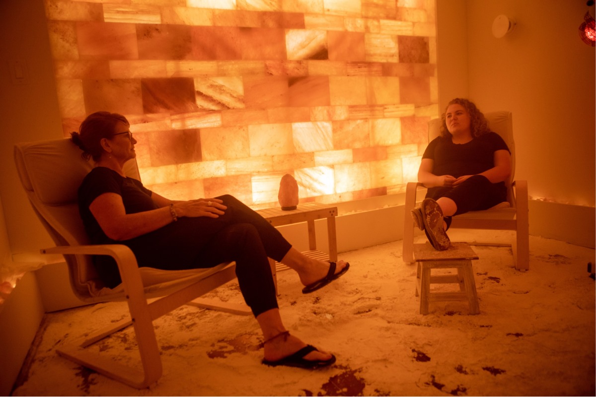 Two Woman Sitting In Chairs On A Salt-Covered Floor In Front Of A Led Backlit Salt Panel Wall At Neurofitness Wellness Center - Ann Arbor, Michigan.