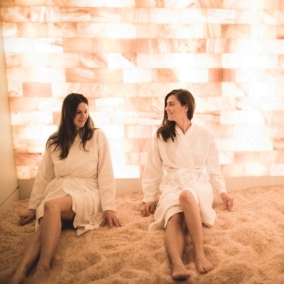 Two Women In White Robes Sitting On A Salt-Covered Floor Against A White Backlit Salt Stone Wall.
