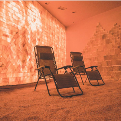 A Salt Cave With Two Reclining Chairs On A Salt-Covered Floor With An Orange Backlit Salt Paneled Wall At The Prana Salt Cave In Wilmington, Nc
