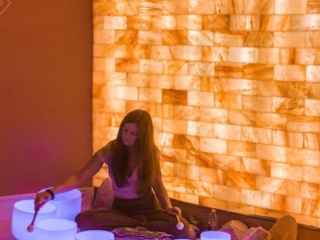 Woman Sitting On A Blanket In Front Of A Led Backlit Salt Panel With White Lit Sound Therapy Bowls At The Prana Salt Cave  - Wilmington, North Carolina.