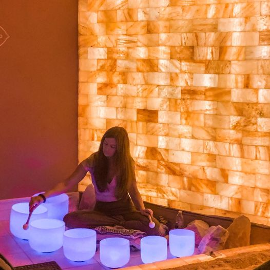 Woman Sitting On A Blanket In Front Of A Led Backlit Salt Panel With White Lit Sound Therapy Bowls At The Prana Salt Cave - Wilmington, North Carolina.