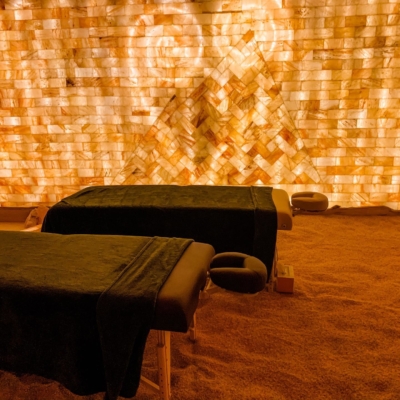 Two Massage Tables On A Salt-Covered Floor In Front Of An Orange Backlit Himalayan Salt Stone Wall.