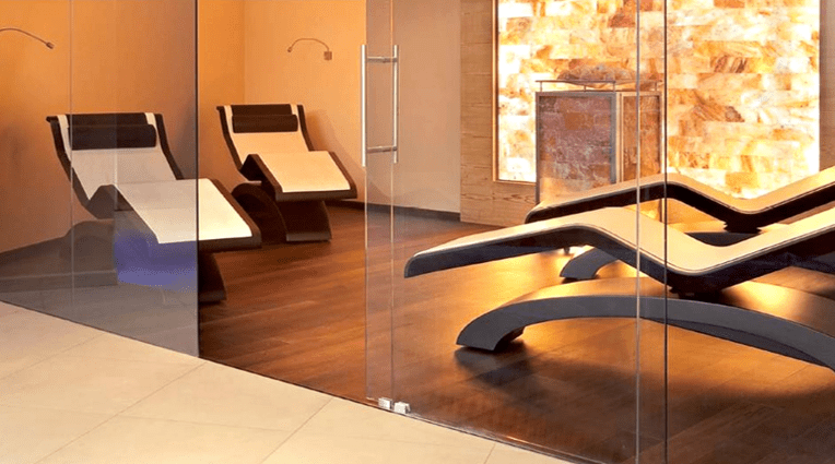 Four white and black chaises on a dark wooden floor through a glass door with an orange backlit Himalayan salt stone wall at a private residence.