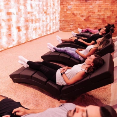 Two Men And Three Women Relaxing On Black Chaises On A Salt-Covered Floor Facing A White Backlit Salt Stone Wall At Pure Salt Studios In Bluffton, South Carolina