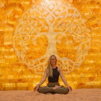 Blonde Haired Women Sitting Cross Legged On A Salt-Covered Floor In Front Of A Dark Yellow Salt Stone Wall With A Tree Made Out Of White Salt As The Wall'S Centerpiece.
