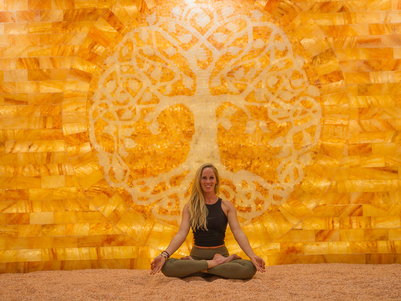 Woman Sitting On A Salt Covered Floor Smiling In Front Of A Led Backlit Salt Panel With A Shape Of A Tree Within At The Purify Wellness Center - Pleasant Grove, Ut.