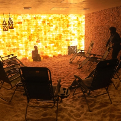 Eight Reclining Chairs On A Salt Covered Floor Surrounded By Himalayan Salt Stone Walls And An Orange Backlit Salt Stone Wall With A Woman Fixing A Chair.