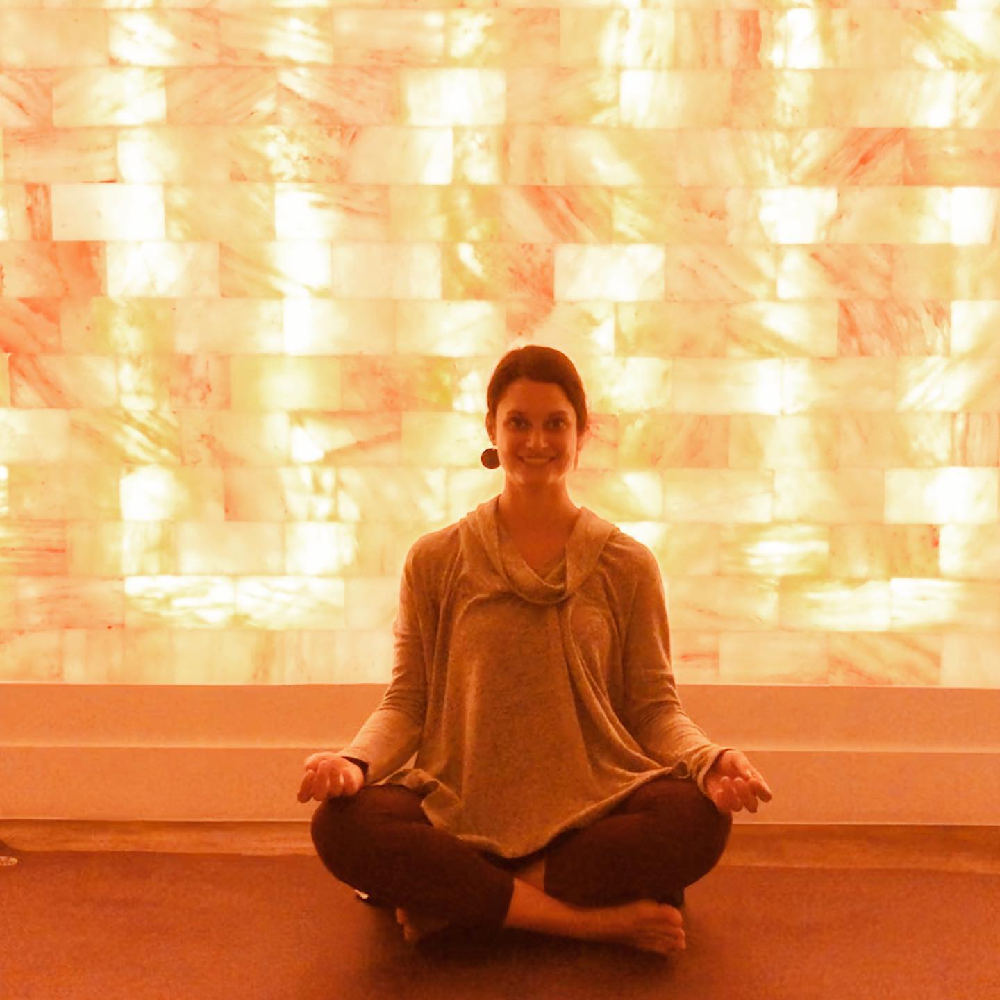 Woman Cross-Legged Smiling In Front Of A White Led Backlit Salt Panel Wall At Sage Blossom Massage - Austin, Texas.