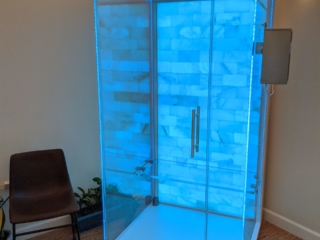 A Blue Backlit Salt Stone Therapy Glass Booth On A Tan Carpet And Brown Chair On The Left At The Sloco Massage And Wellness Spa