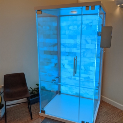 A blue backlit salt stone therapy glass booth on a tan carpet and brown chair on the left at the Sloco Massage and Wellness Spa