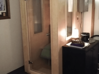 A Wooden And Glass Salt Booth With A Black Cabinet On The Floor With Decor On A Dark Grey Rug With A Tan And White Foot Mat At The Entry Of The Booth.