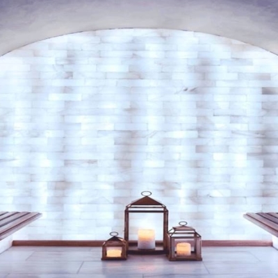 A White Backlit Salt Stone Wall With Three Lit Candles In Wooden Containers And Two Dark Wooden Benches.
