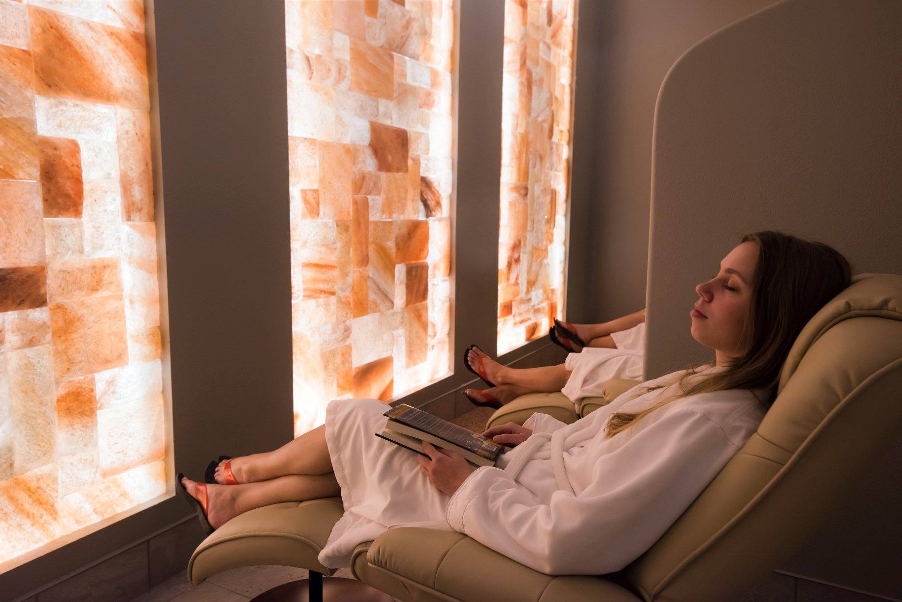Woman In A White Robe Relaxing In A Lounge Chair Facing A Led Backlit Salt Panel Wall At The Sundara Inn And Spa - Wisconsin Dells, Wisconsin.