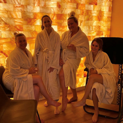 Four Women, Two Standing And Two Sitting, Smiling In Front Of An Orange Backlit Himalayan Salt Stone Wall In A Halotherapy Room.
