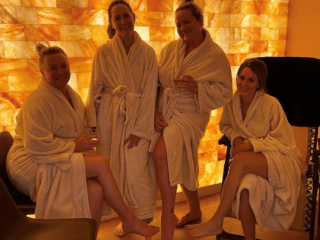 Four Women In Spa Robes In A Dimly Lit Salt Room With An Led Backlit Salt Brick Wall