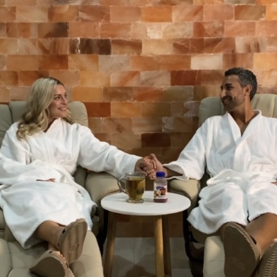 Women And Man Sitting In Lounge Chairs Both In White Robes Holding Hands In Front Of A Himalayan Salt Panel Wall At The Salt Center - Alpharetta, Georgia.