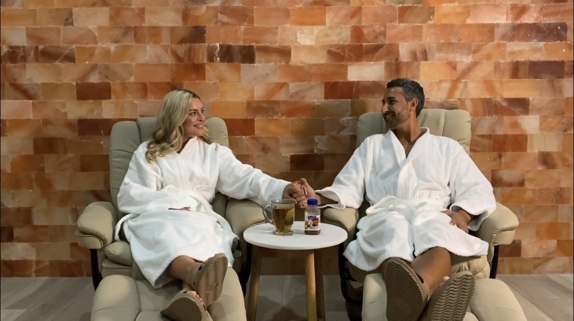 Women And Man Sitting In Lounge Chairs Both In White Robes Holding Hands In Front Of A Himalayan Salt Panel Wall At The Salt Center - Alpharetta, Georgia.
