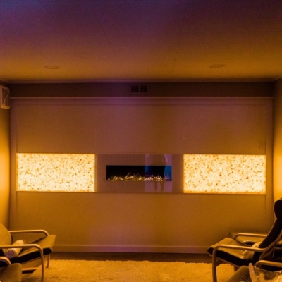 Four Grey Chairs In A Salt Therapy Room On A Salt-Covered Floor With An Electric Fireplace On The Wall Centered By Two Orange Backlit Salt Stone Panels.