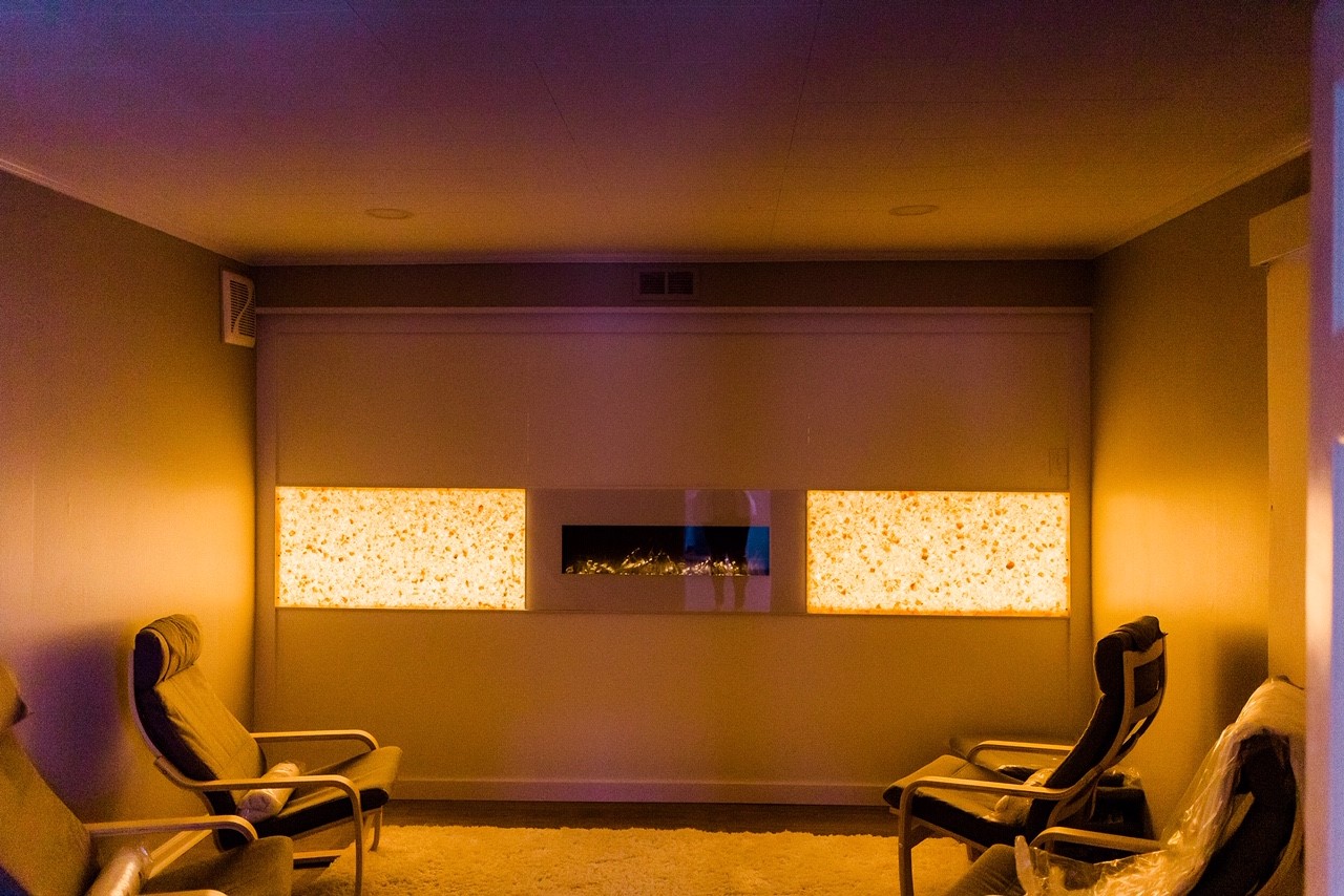Four Grey Chairs In A Salt Therapy Room On A Salt-Covered Floor With An Electric Fireplace On The Wall Centered By Two Orange Backlit Salt Stone Panels.