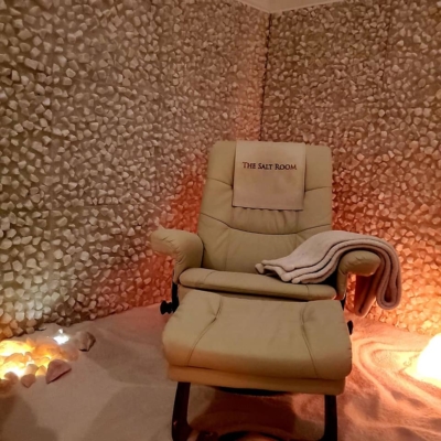A White Chair In A Salt Therapy Room With A White Blanket On A White Salt-Covered Floor Surrounded By Salt-Stone Covered Walls And Dim Ambient Orange Lighting At The Salt Room Halifax Nova Scotia.