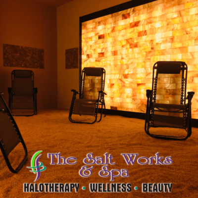 Three Reclining Chairs With A Salt Stone Paneled Wall Backlit With Orange Lighting With Words That Say &Quot;The Salt Works &Amp; Spa Halotherapy - Wellness - Beauty&Quot; At The Bottom.