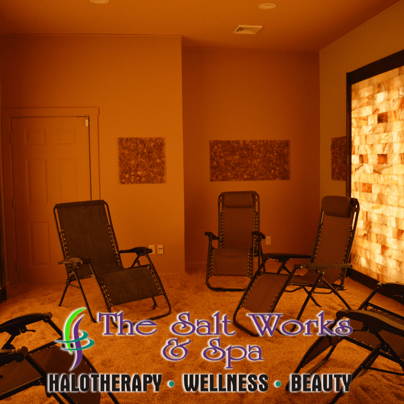 Five Reclined Chairs In A Salt Chamber With A Backlit Orange Salt Paneled Wall With Words Saying &Quot;The Salt Works &Amp; Spa Halotherapy - Wellness - Beauty&Quot; At The Bottom.