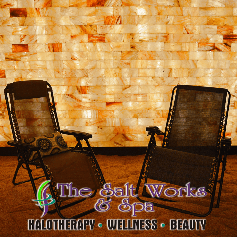 Two Reclining Chairs On A Salt-Covered Floor One With A Pillow In Front On A Salt Stone Wall Backlit By Orange Lighting With The Saying &Quot;The Salt Works &Amp; Spa Halotherapy - Wellness - Beauty&Quot;  On The Bottom Of The Picture.