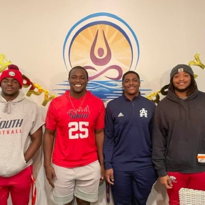 Football players from University of South Alabama smiling in front of a tan wall at Above and Beyond Yoga and Salt Therapy.