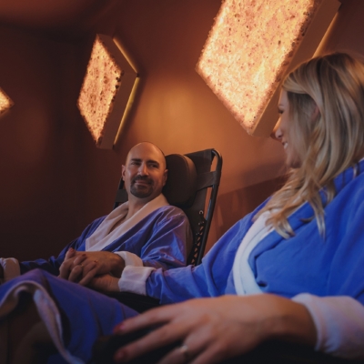 One Woman And One Man In Blue Robes Holding Hands Smiling At Each Other In A Salt Therapy Room With Diamond Salt Stone Wall Décor At The Unwind Westclox Peru Ilinois