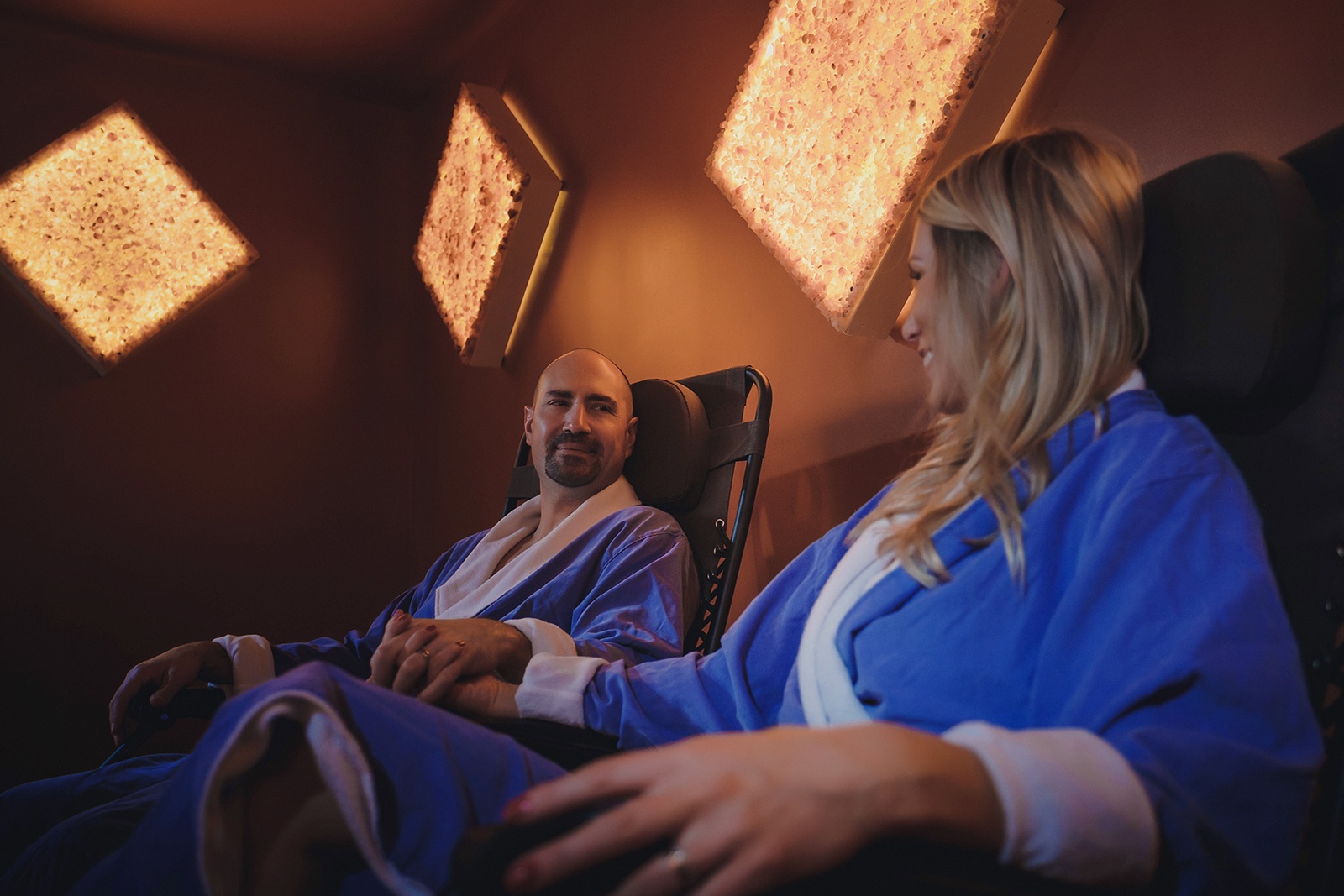 One Woman And One Man In Blue Robes Holding Hands Smiling At Each Other In A Salt Therapy Room With Diamond Salt Stone Wall Décor At The Unwind Westclox Peru Ilinois