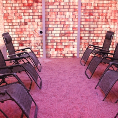 Seven Grey Reclining Chairs On A Himalayan Salt-Covered Floor Surrounded By Two Salt Stone Walls Backlit By Pinkish Lighting And A Salt Paneled Wall Back Lit By Pinkish Lighting At The Urban Oasis Salt Spa In Traverse City Michigan