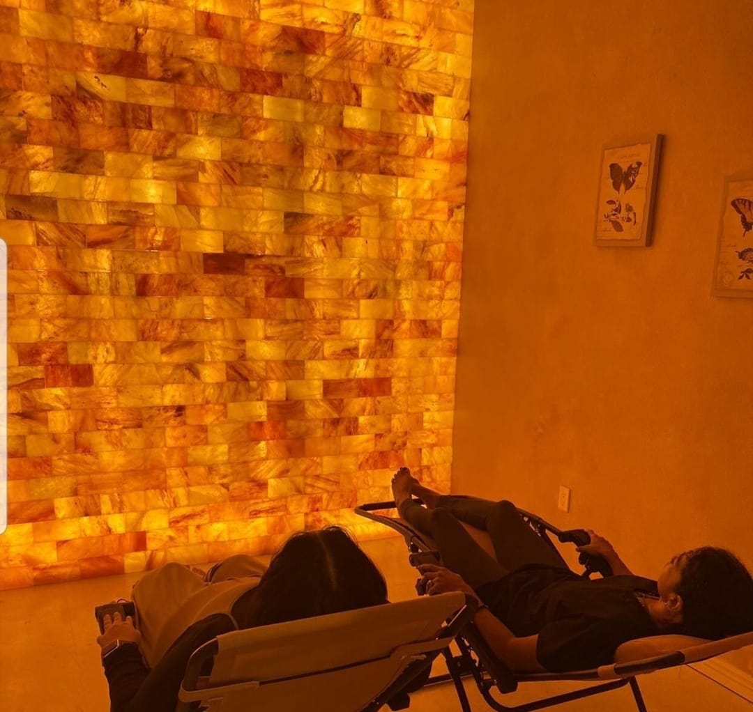 Two Women Reclined In Chairs Relaxing In Front Of A Led Backlit Salt Panel Wall With Two Butterfly Pictures On The Wall At Venus Moon Wellness Boutique - Maricopa, Arizona.