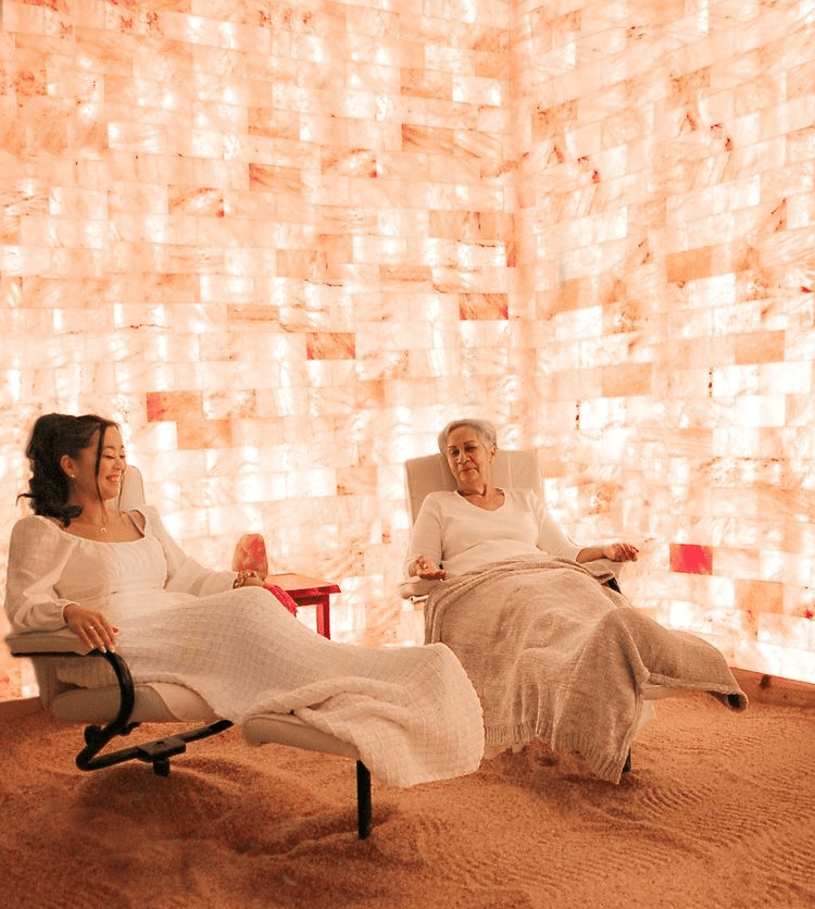 Two Women Relaxing In Reclined Chairs On A Salt-Covered Floor Surrounded By White Led Backlit Salt Panels At Wellcome Om Integral Healing &Amp; Education Center - Spring Hill, Florida.