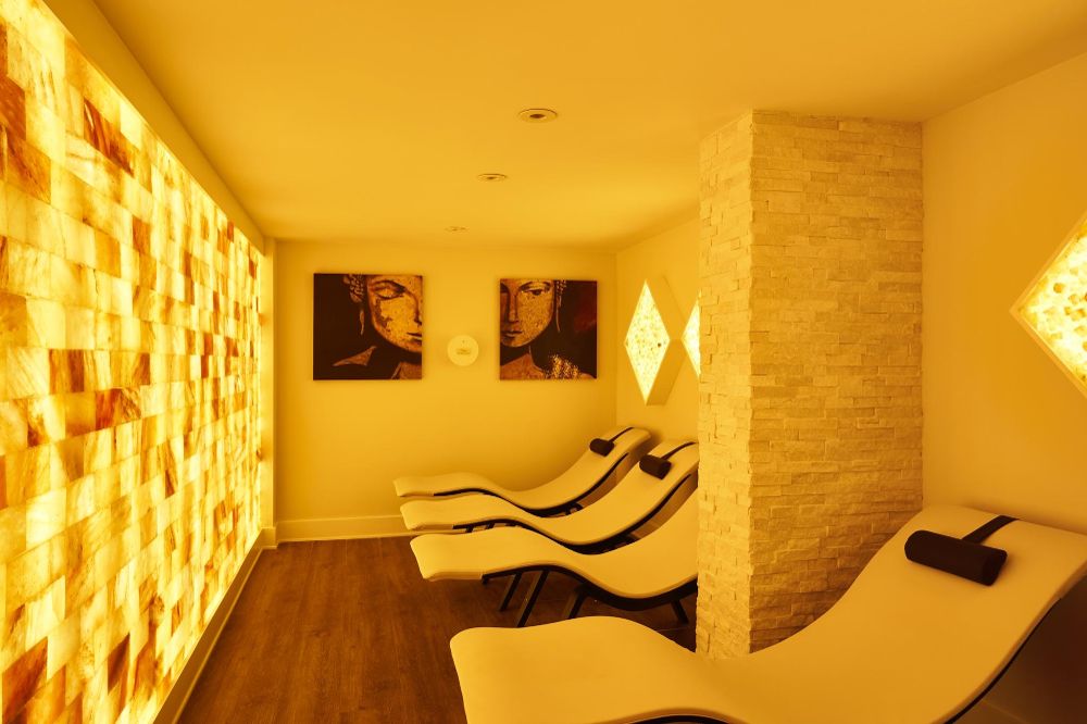 Four White Lounge Chairs With Brown Headrests With A Salt Paneled Wall Backlit By Yellow Lighting, Three Diamond Shaped Salt Stone Décor, And Two Darkish Red Pieces Of Art With Faces At The White Orchid Spa In Vero Beach.